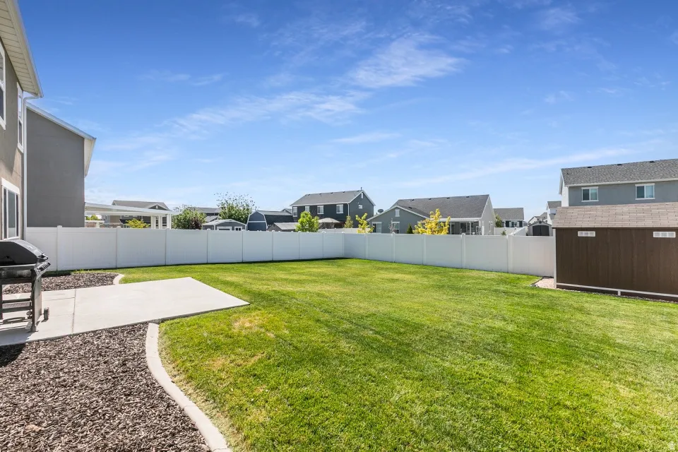 Expansive grassy yard with a white privacy fence, concrete patio, and a storage shed with wood-look siding