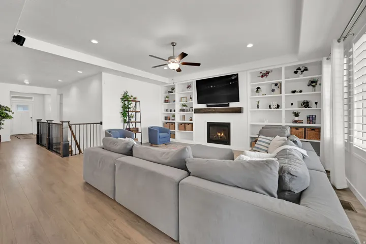Living area featuring wood-finish flooring, a built-in fireplace, extensive built-in shelving, a ceiling fan, and large window with plantation shutters