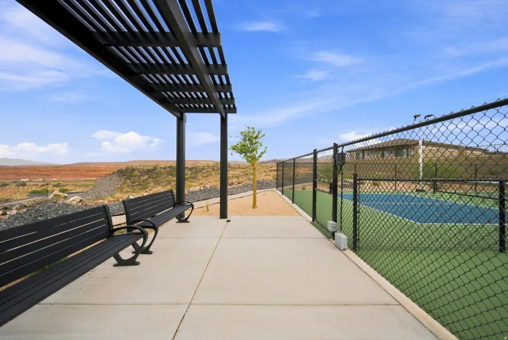 Pergola-covered seating area with concrete paving, adjacent to a tennis court featuring chain-link fencing