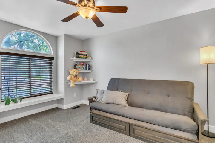 Arch-top window with blinds, ceiling fan with light fixture, built-in corner shelving, and gray carpet flooring