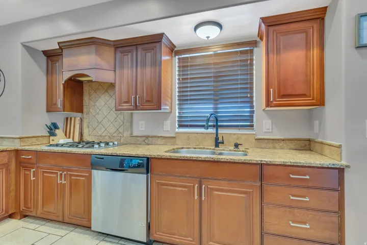 Warm wood-finish cabinetry with silver hardware, light-colored stone countertops, a tile backsplash, and a stainless steel dishwasher