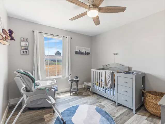 Wood-finish flooring, ceiling fan with integrated lighting, window with white curtains, and light gray wall paint