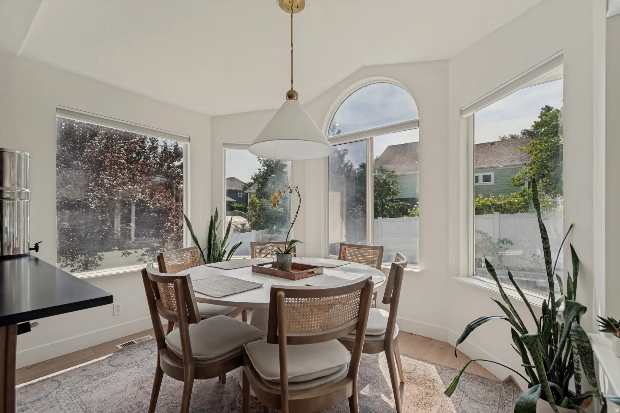 Bright dining space showcasing a curved arched window with expansive natural light, wood-frame dining chairs, neutral wood-finish flooring, and a pendant light fixture