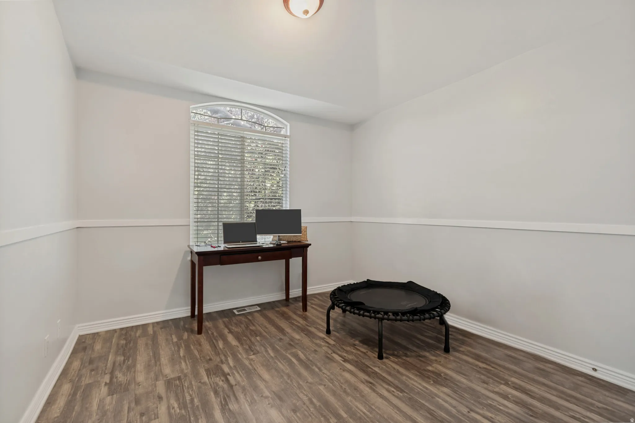 Versatile office or studio space featuring wood-finish flooring, clean white walls with chair rail molding, a large window with plantation shutters providing natural light, and recessed ceiling lighting