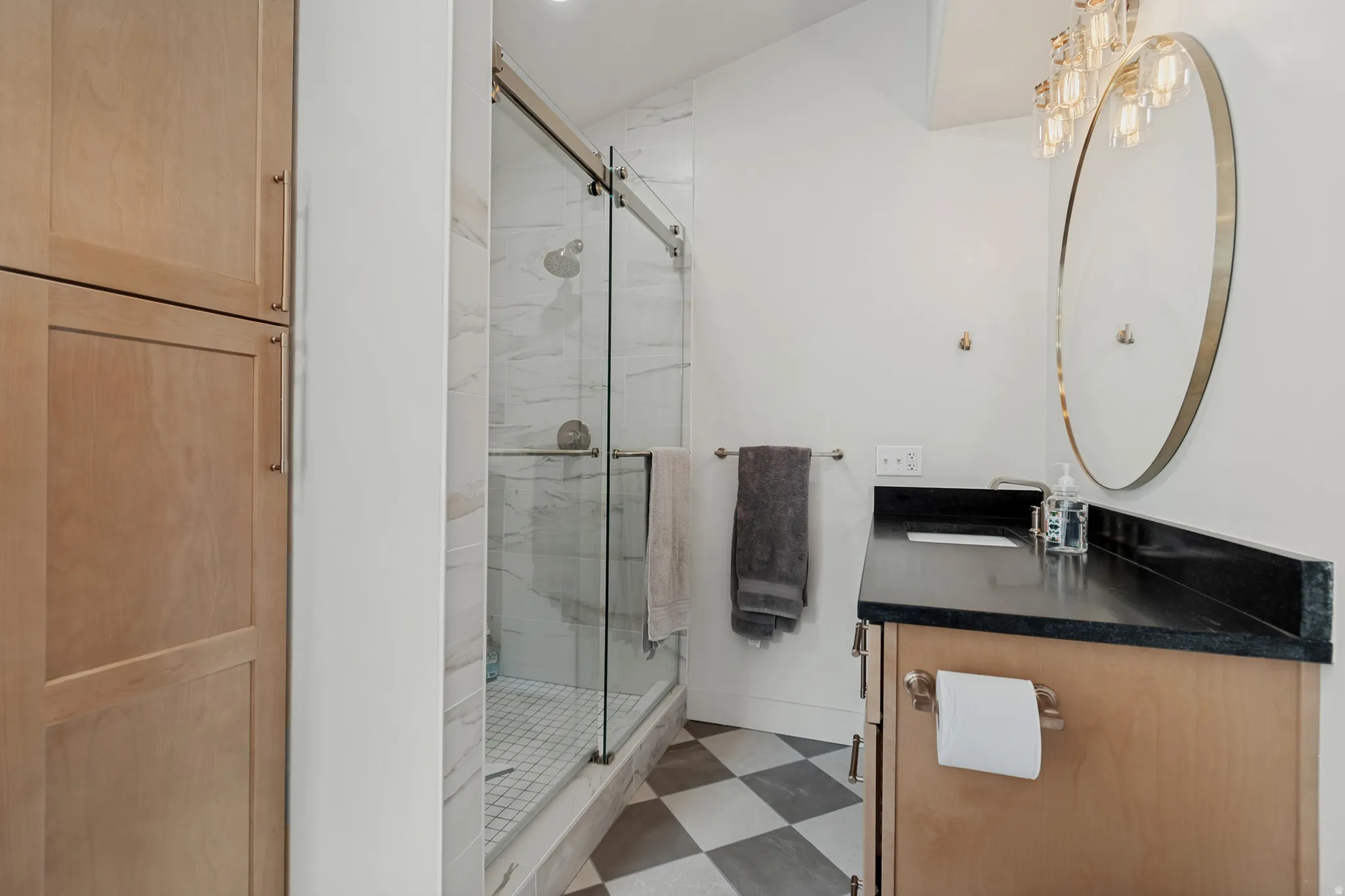Modern bathroom featuring a walk-in shower with glass enclosure and tile surround, black countertop with integrated sink, circular mirror with brushed metal frame, wood-finish cabinetry, and geometric tile flooring in neutral tones