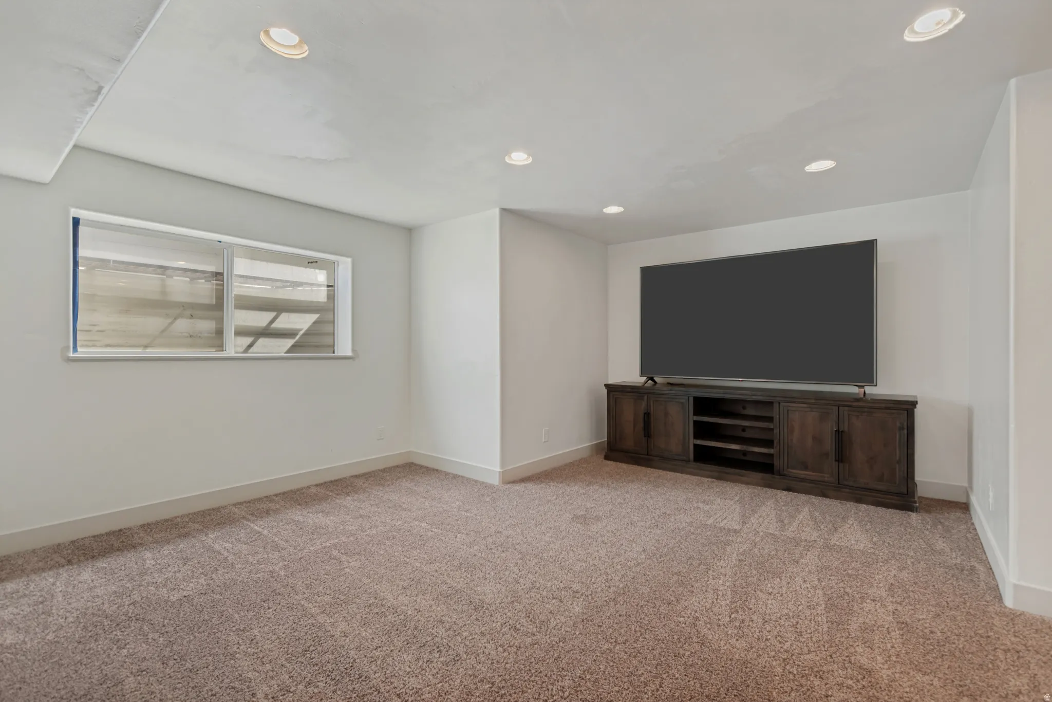 Spacious recreation room featuring plush carpeting, recessed ceiling lighting, and a custom wood-finish media console with integrated shelving