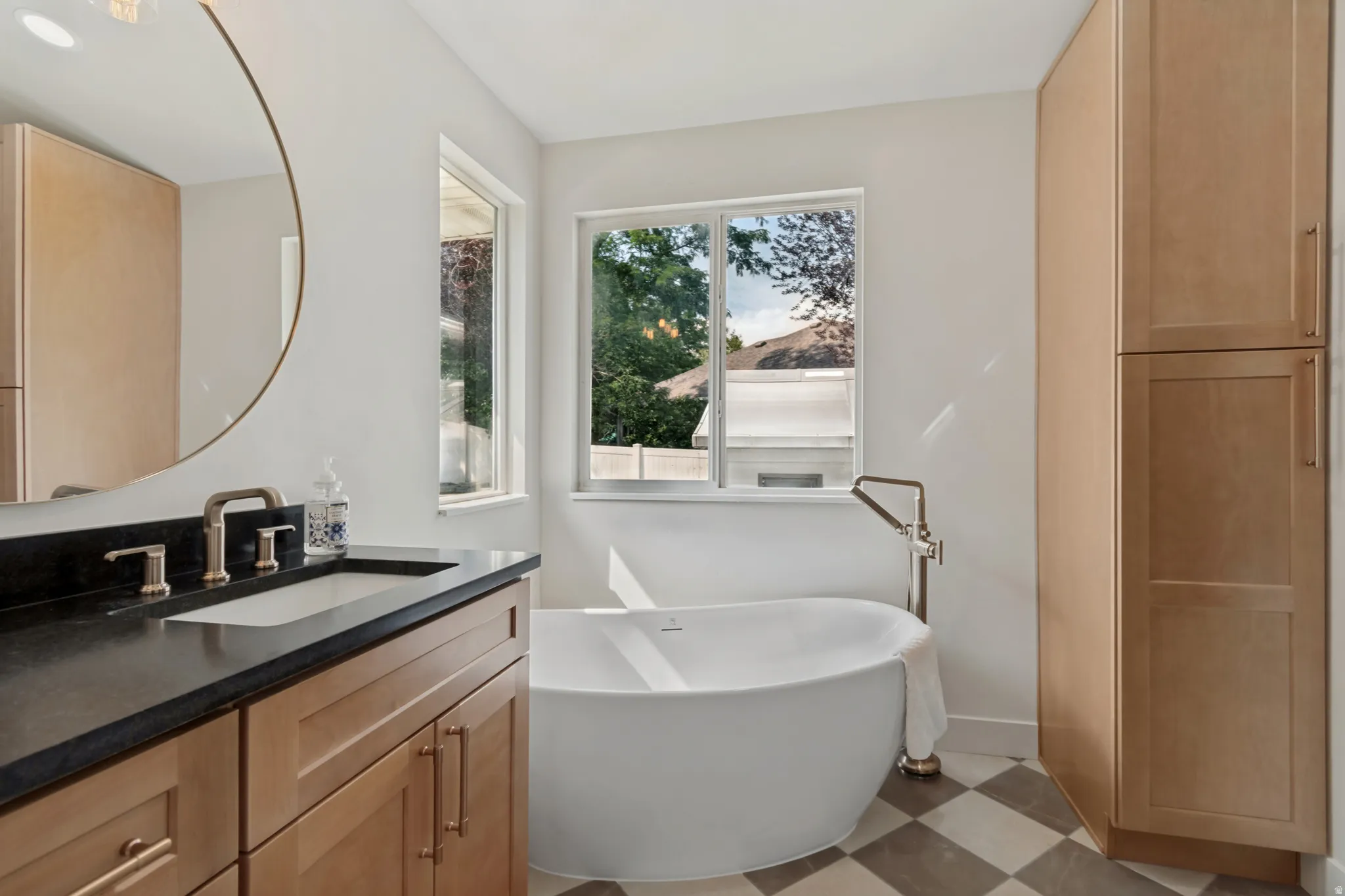Freestanding soaking tub with floor-mounted faucet, dual windows, wood-finish cabinetry, and black and white checkerboard flooring