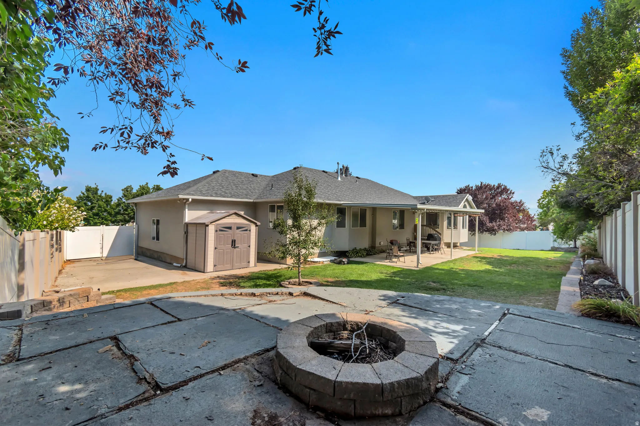 Expansive backyard oasis featuring a circular fire pit with stone surround, mature landscaping with established trees, manicured lawn, and generous hardscape patio space