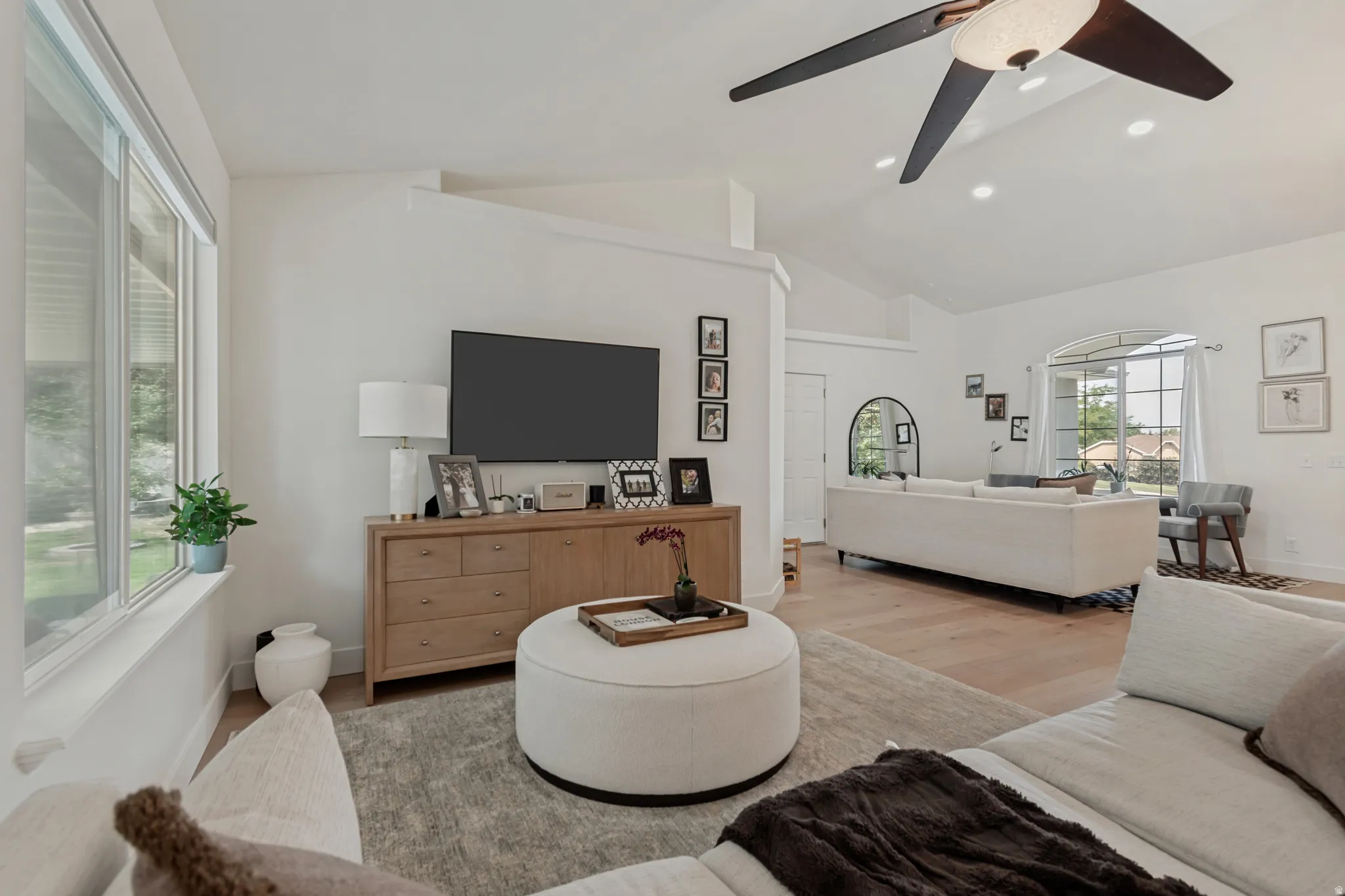 Bright, open-concept living area featuring a wood-finish entertainment console with integrated media display, a ceiling fan with modern blade design, large windows with natural light exposure, light wood-finish flooring throughout, and a curved upholstere