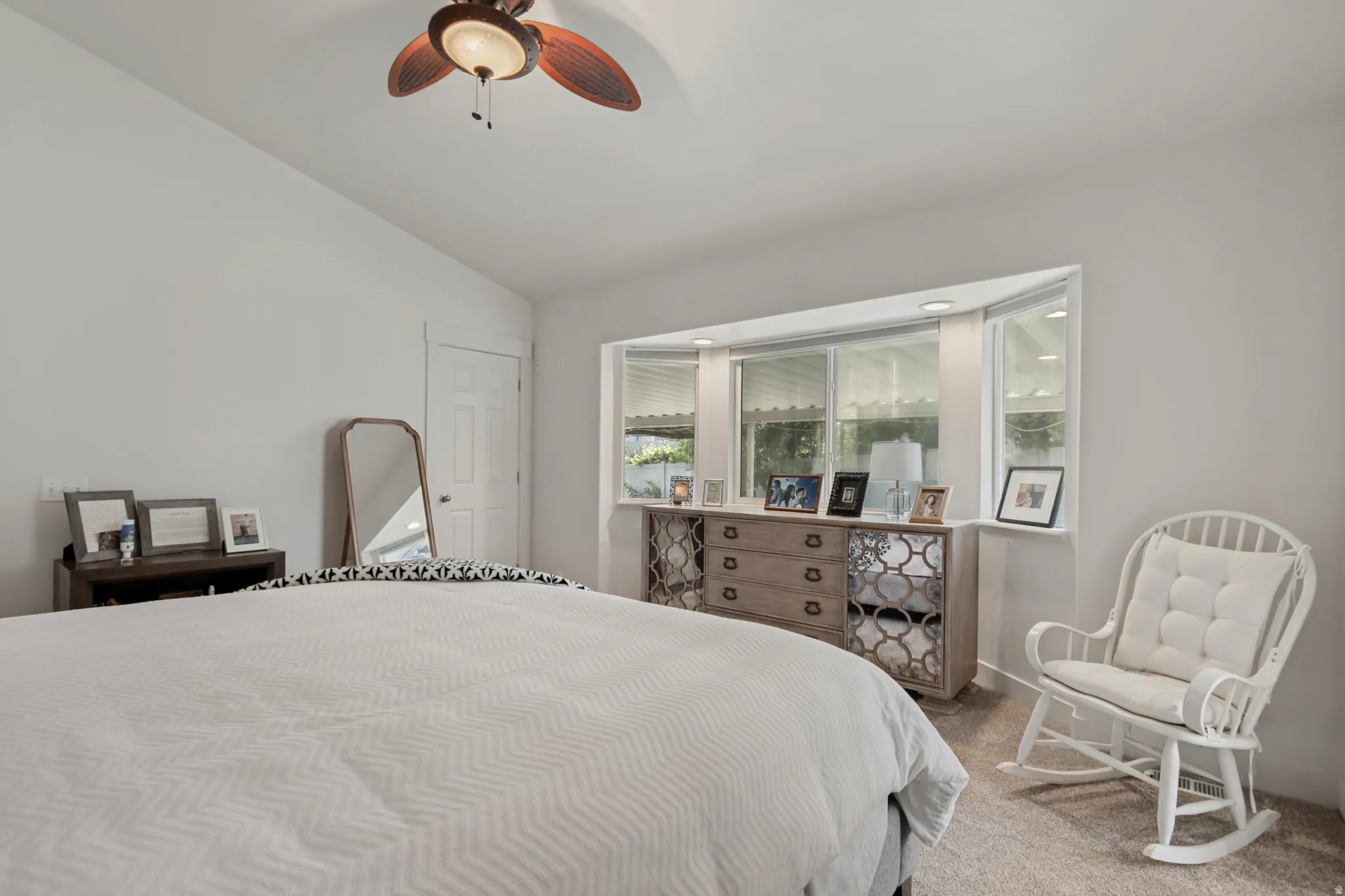 Serene primary bedroom showcasing neutral carpeting, ceiling fan with integrated lighting, mirrored dresser with decorative hardware, and glass sliding doors opening to a private balcony with outdoor views