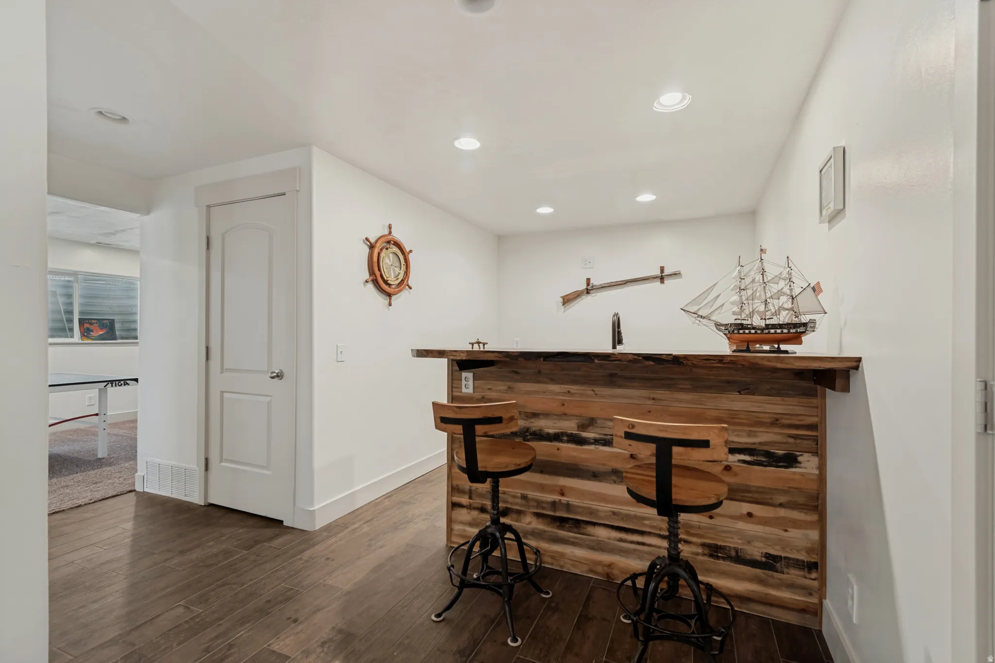 Nautical-themed bar area featuring rich wood cabinetry with black metal accents, recessed ceiling lighting, ship's wheel wall décor, and dark wood-finish flooring throughout the space