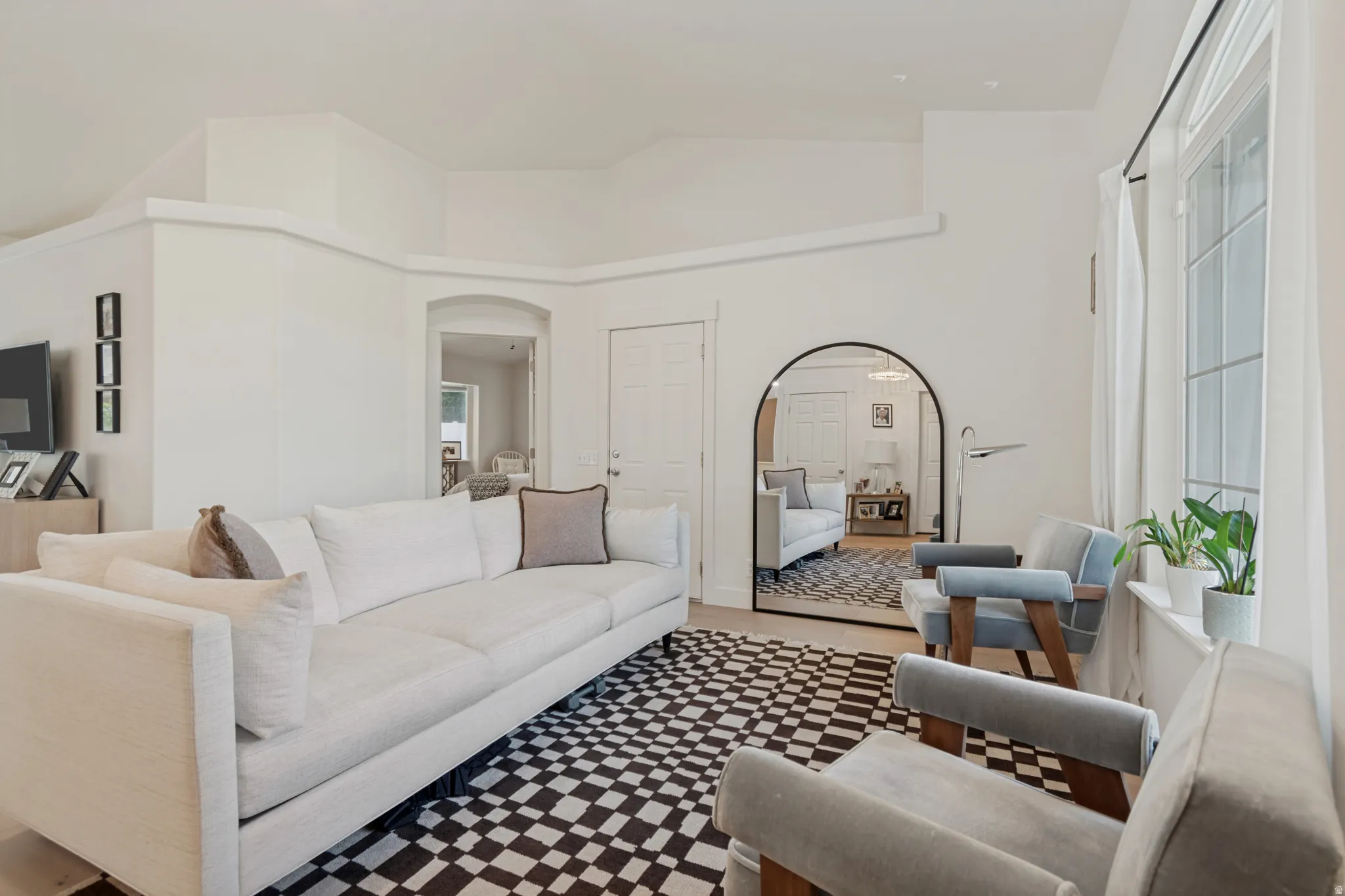 Sophisticated living room featuring a curved white sectional sofa, geometric patterned hardwood flooring, arched mirror with bronze-finish frame, floor-to-ceiling windows with natural light, and neutral wall finishes throughout the open-concept space
