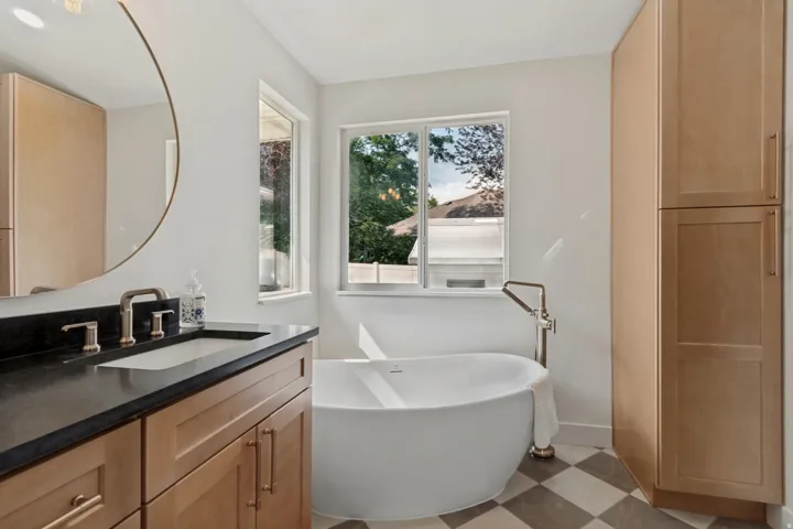 Freestanding soaking tub with floor-mounted faucet, dual windows, wood-finish cabinetry, and black and white checkerboard flooring