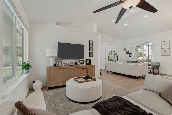 Bright, open-concept living area featuring a wood-finish entertainment console with integrated media display, a ceiling fan with modern blade design, large windows with natural light exposure, light wood-finish flooring throughout, and a curved upholstere