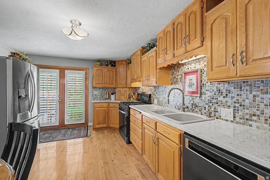 Wood-finish flooring, light wood cabinetry, dual basin sink with a gooseneck faucet, light-toned countertops, and a full tile backsplash