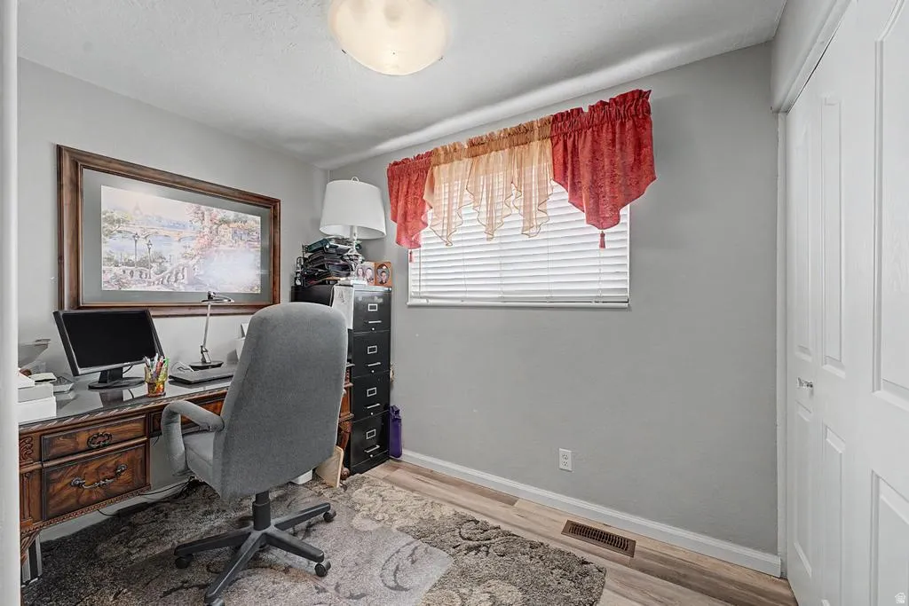 Dedicated home office featuring soft gray walls, wood-finish flooring, and a large window with red valance and horizontal blinds