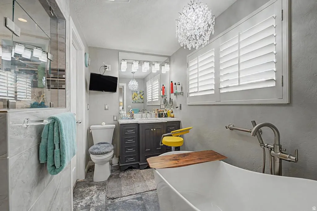Freestanding soaking tub with floor-mounted faucet, plantation shutters, crystal chandelier, vanity with dual sinks, and walk-in shower with glass enclosure