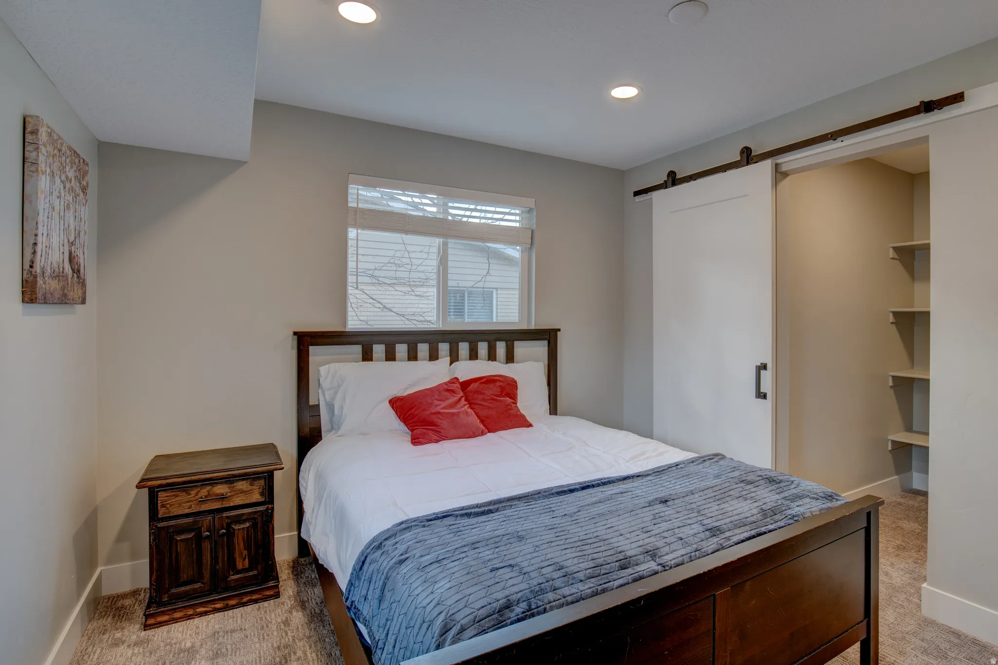 Spacious bedroom featuring a wood-frame bed, recessed ceiling lighting, large double-hung windows with cellular shades, carpeted flooring, and a walk-in closet with built-in shelving and barn-style sliding door hardware