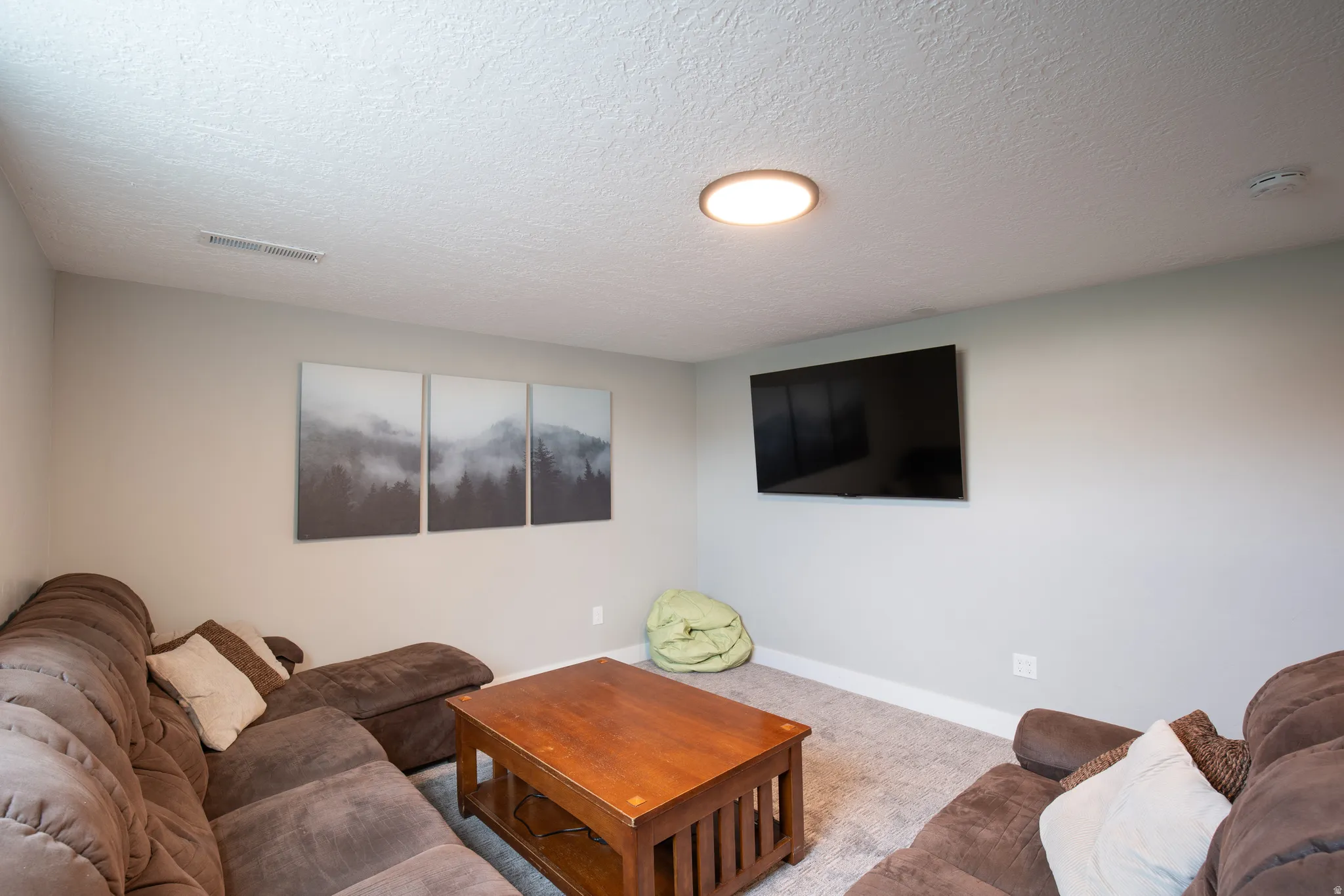Finished basement recreation room featuring plush seating arrangements, wood-finish coffee table, recessed ceiling lighting, wall-mounted flat-screen television, and neutral carpeting throughout