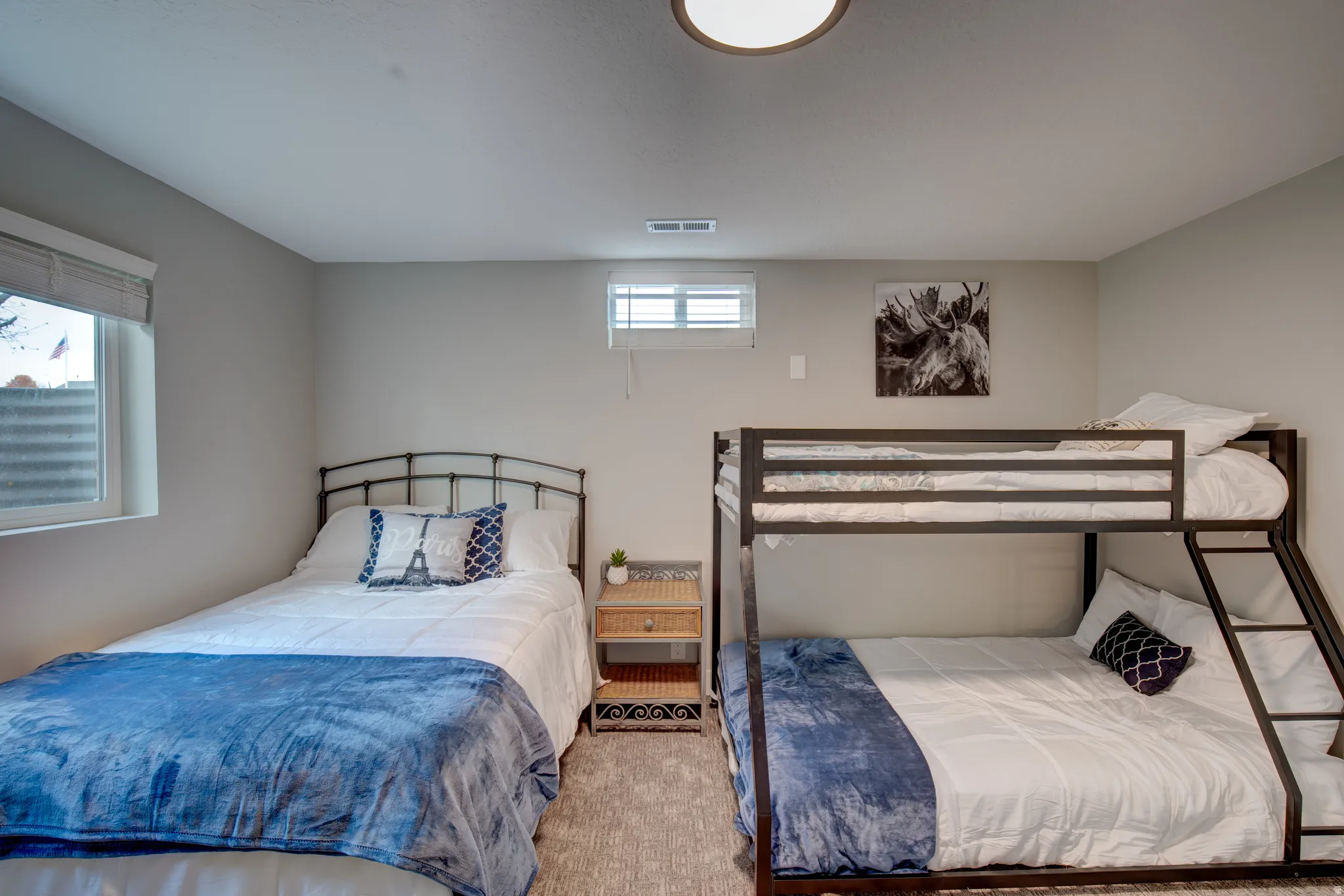 Carpeted room with a ceiling-mounted light fixture, two windows with blinds, a metal-frame bed, and a metal-frame bunk bed