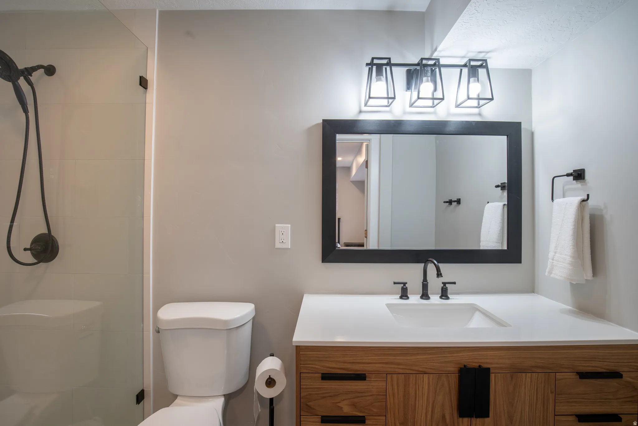 Glass-enclosed shower with dark bronze fixtures, a wall-mounted toilet, wood-finish vanity with an integrated sink, and dark bronze wall-mounted faucet