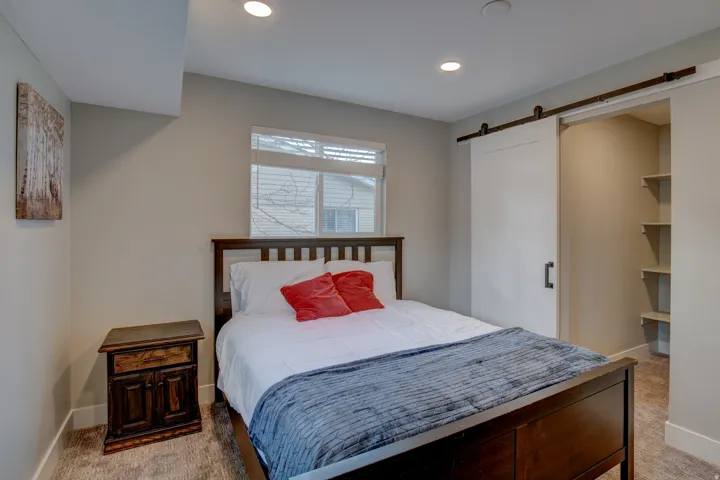 Spacious bedroom featuring a wood-frame bed, recessed ceiling lighting, large double-hung windows with cellular shades, carpeted flooring, and a walk-in closet with built-in shelving and barn-style sliding door hardware