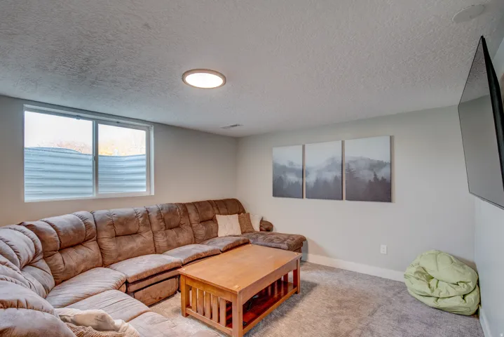 Comfortable recreation room featuring a spacious sectional sofa, wood-finish coffee table, neutral carpet flooring, recessed ceiling lighting, and large windows with horizontal blinds allowing natural light throughout the space