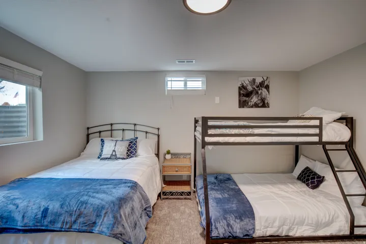 Carpeted room with a ceiling-mounted light fixture, two windows with blinds, a metal-frame bed, and a metal-frame bunk bed