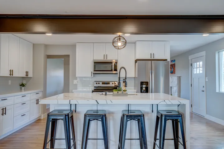 Modern kitchen featuring an extended island with a waterfall edge, stainless steel appliances, white flat-panel cabinetry, and wood-finish flooring