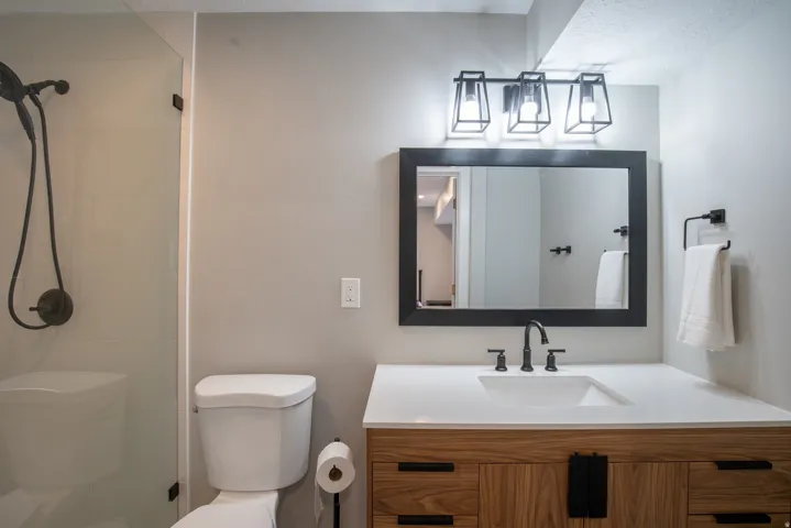 Glass-enclosed shower with dark bronze fixtures, a wall-mounted toilet, wood-finish vanity with an integrated sink, and dark bronze wall-mounted faucet