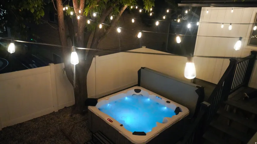 Integrated hot tub with illuminated water, string lighting, white privacy fencing, and wood-finish decking