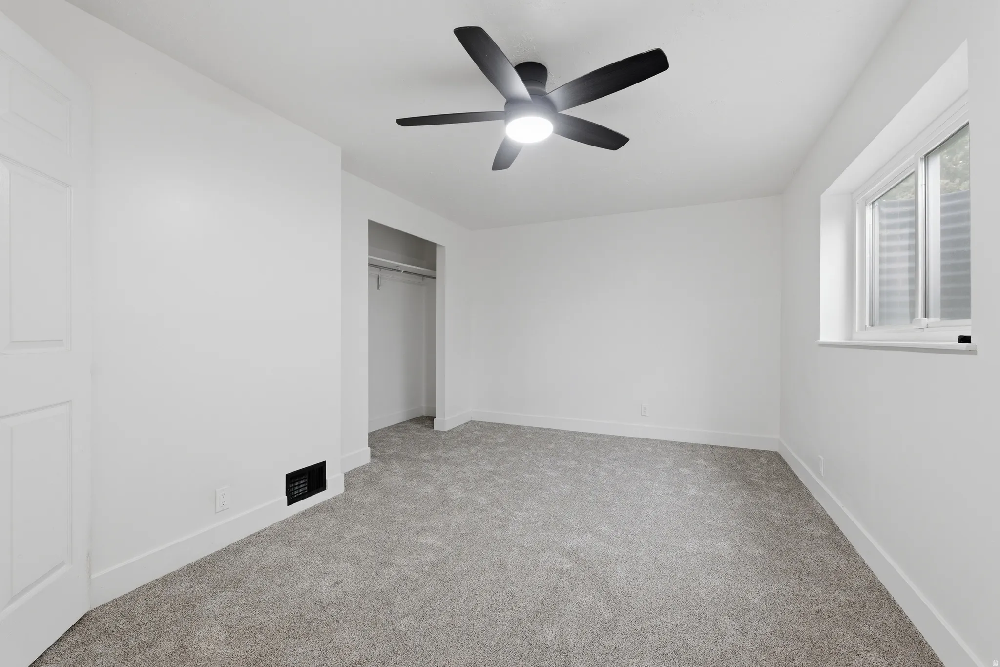 Wall-to-wall carpet flooring, white painted walls, a ceiling fan with integrated lighting, a window with a white frame, and a built-in closet with a hanging rod