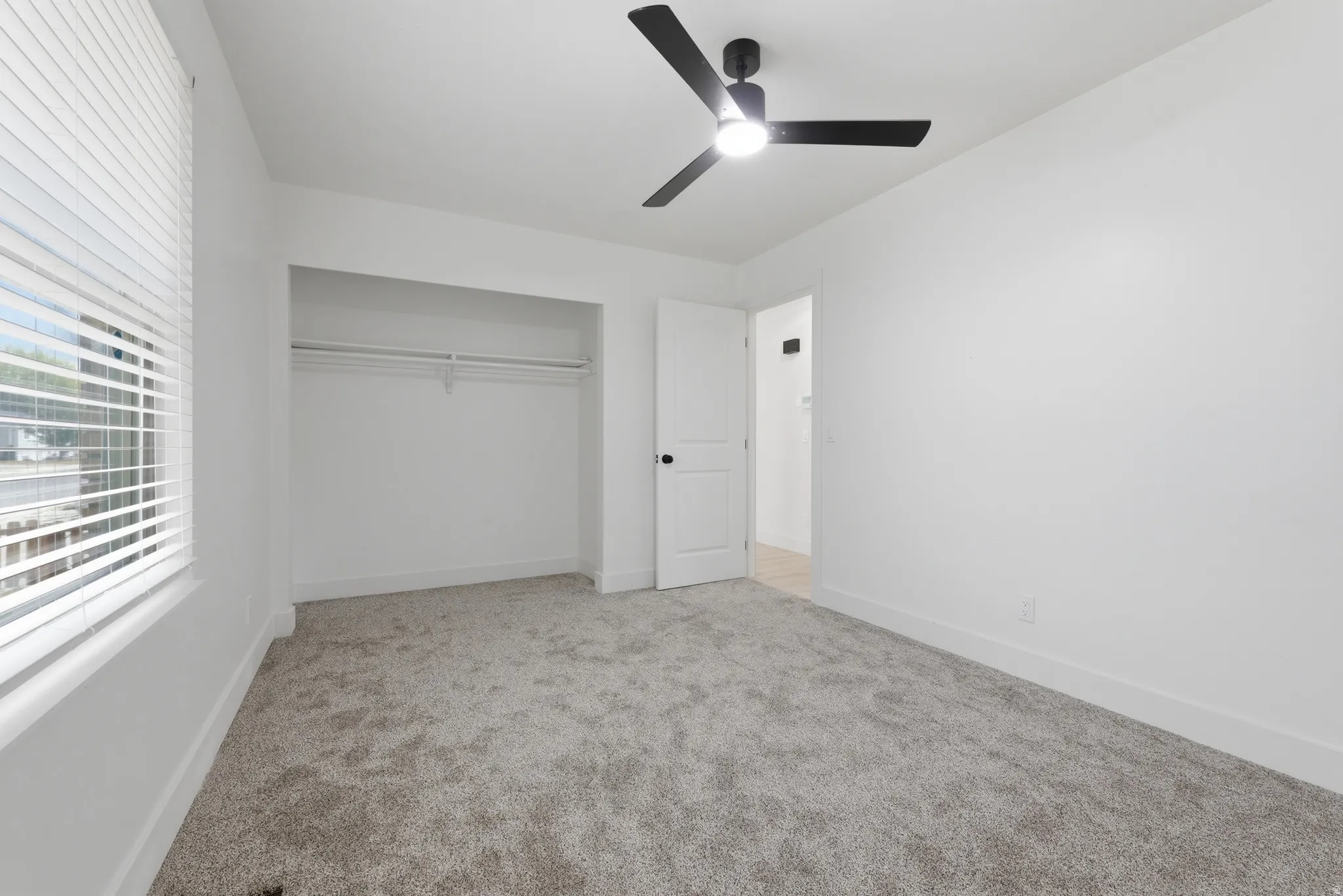 Recessed closet with shelving, modern ceiling fan with light, window with horizontal blinds, neutral-toned carpeting, and white interior door with black hardware