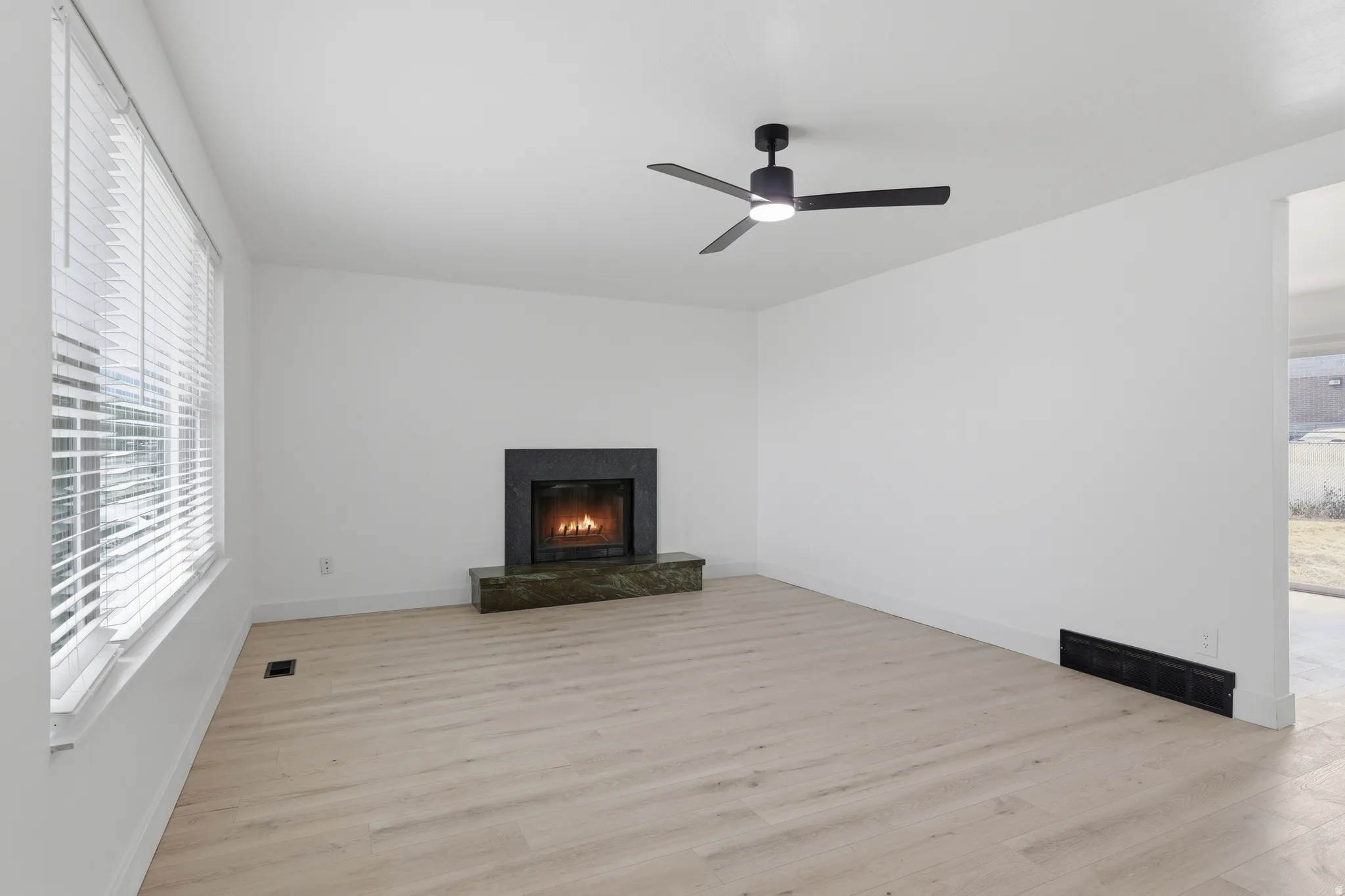 Light wood-finish flooring, a fireplace with a dark stone surround and hearth, a ceiling fan with integrated lighting, and white window blinds
