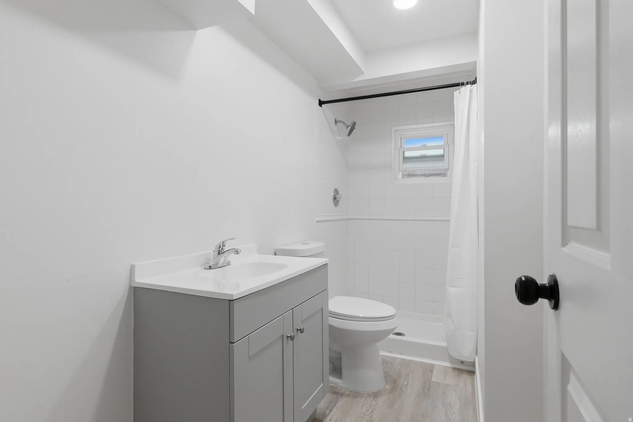 Vanity with an integrated sink and chrome faucet, wood-finish flooring, white subway tile shower surround, and a compact window