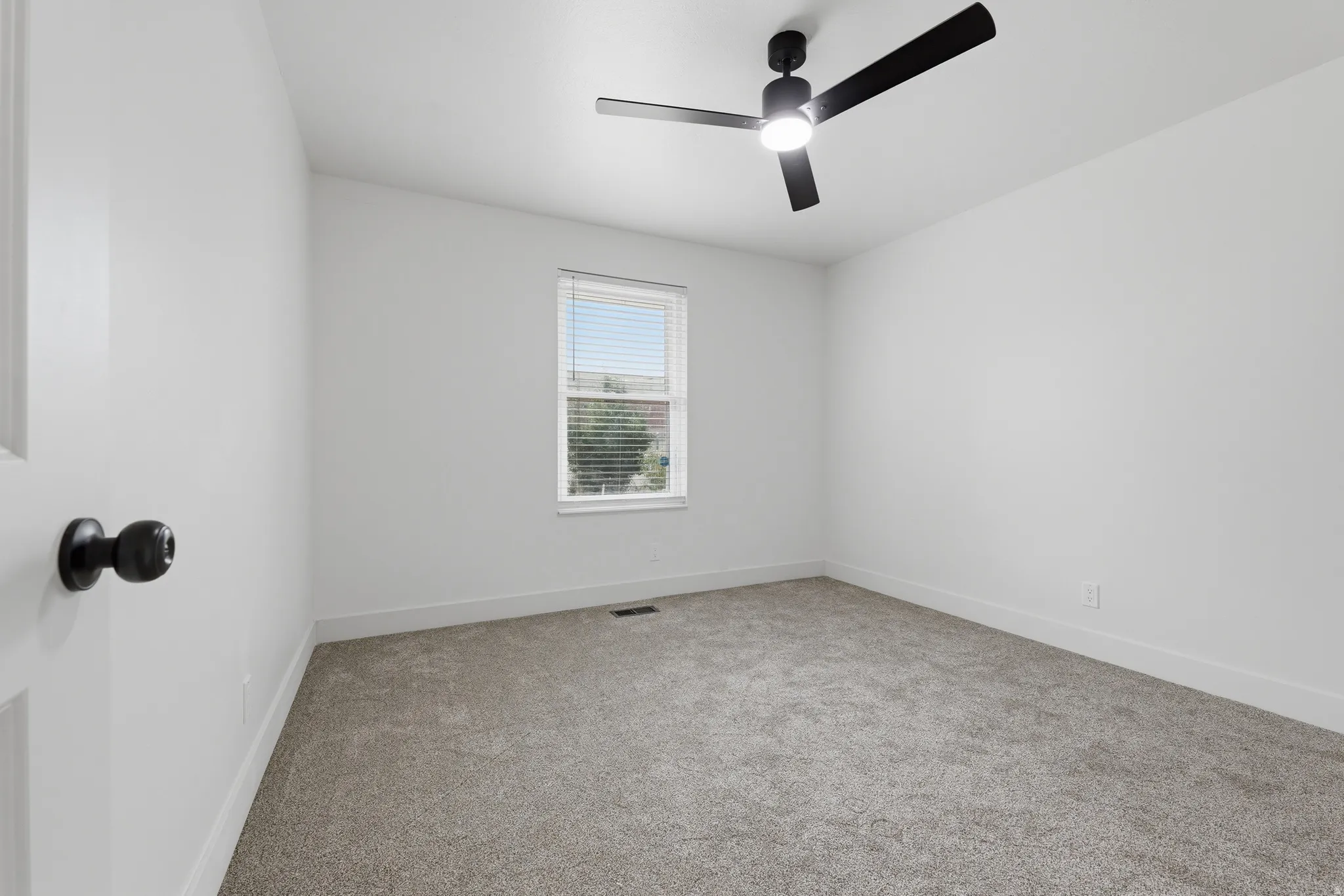White walls with gray carpeting, a single window with blinds, and a black ceiling fan with an integrated light