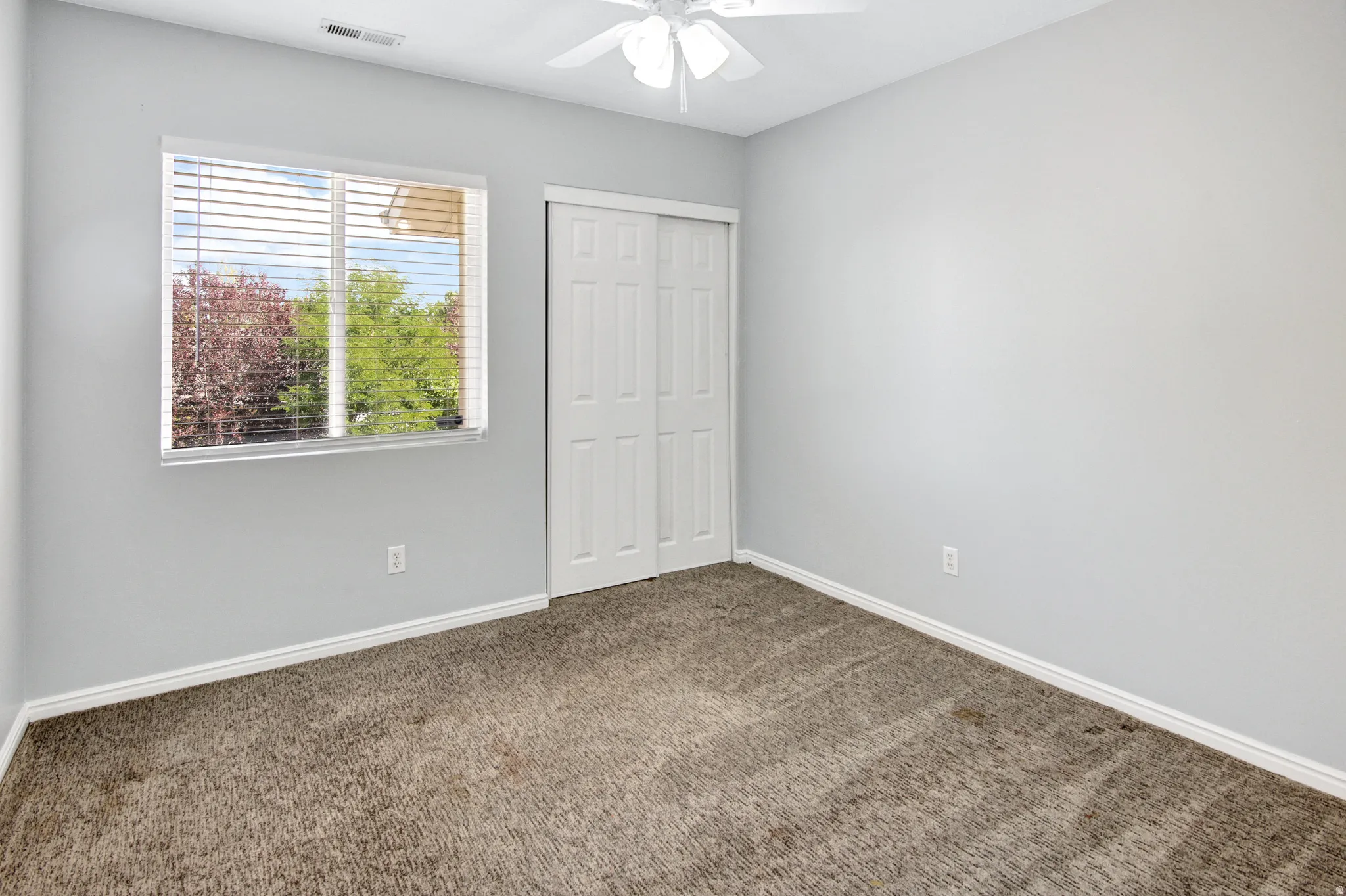 Neutral wall color, carpeted flooring, sliding closet doors, window with horizontal blinds, and a ceiling fan with integrated lighting