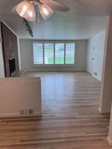 Wood-finish flooring throughout, brick fireplace with mantel, window with plantation shutters, and ceiling fan with light fixture