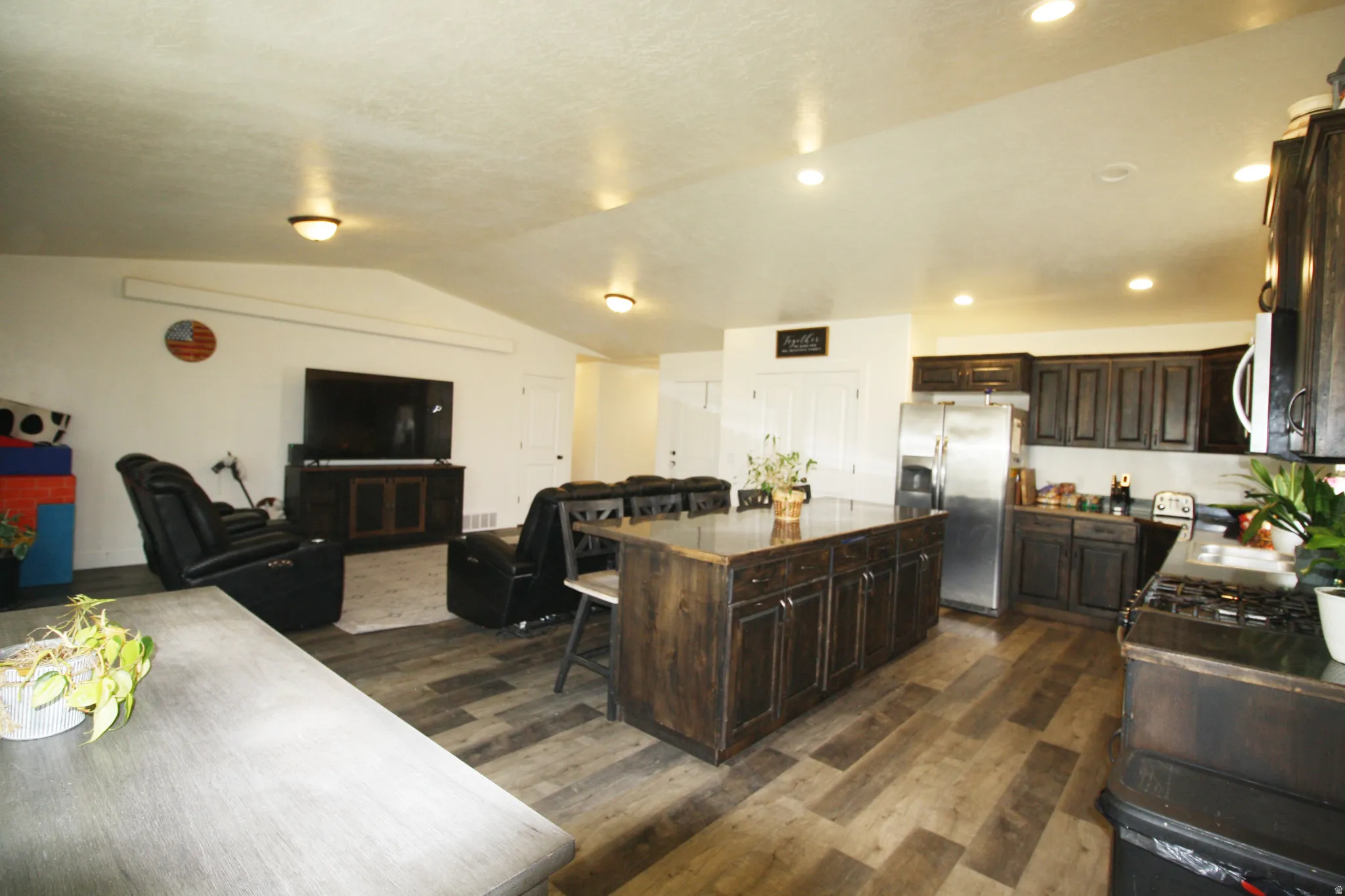 Spacious open-concept layout featuring wood-finish flooring, dark wood cabinetry, a large kitchen island, stainless steel refrigerator, and recessed lighting