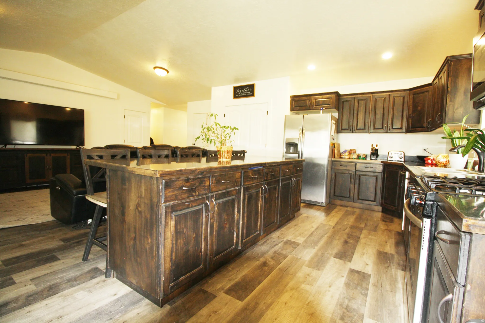 Spacious kitchen featuring an expansive center island, wood-finish flooring, dark wood cabinetry, stainless steel refrigerator, and a gas range