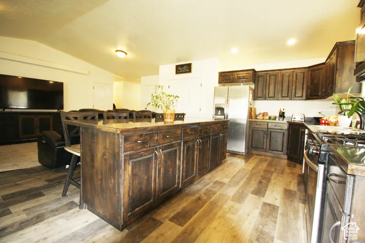 Spacious kitchen featuring an expansive center island, wood-finish flooring, dark wood cabinetry, stainless steel refrigerator, and a gas range
