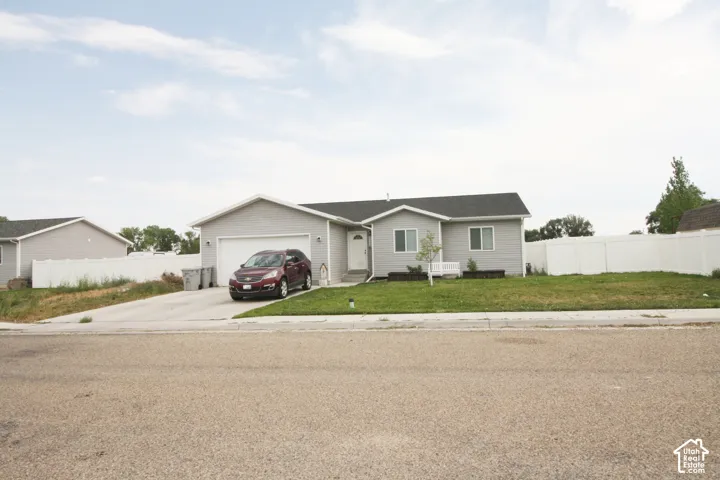 Single-story residence featuring light gray siding, an attached garage, a paved driveway, a green lawn, and a white privacy fence