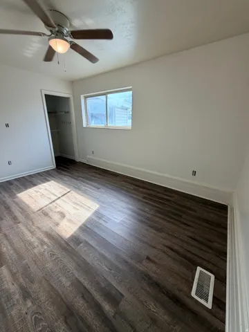 Spacious room with dark wood-finish flooring, ceiling fan with integrated lighting, large window with natural light, and white painted walls with electrical outlets throughout