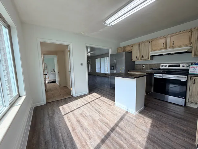 Spacious kitchen featuring wood-finish cabinetry, stainless steel appliances including dual wall ovens, a central island with white finish, black countertops, and abundant natural light from large windows