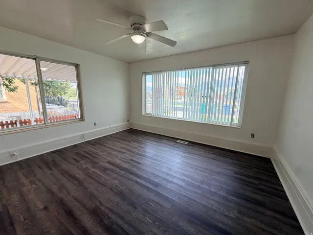 Spacious room featuring wood-finish flooring, white walls, multiple large windows with blinds, and a ceiling fan with integrated lighting