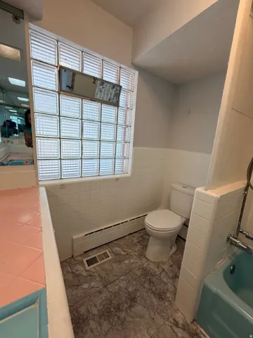 Glass block window, white tile wainscoting, light-colored walls, toilet, and a bathtub with fixtures