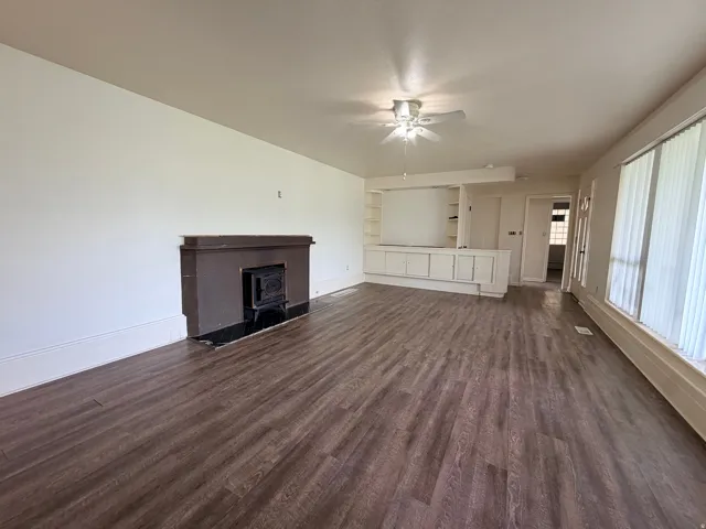 Wood-finish flooring, built-in shelving, fireplace with black hearth, ceiling fan, and large windows with vertical blinds