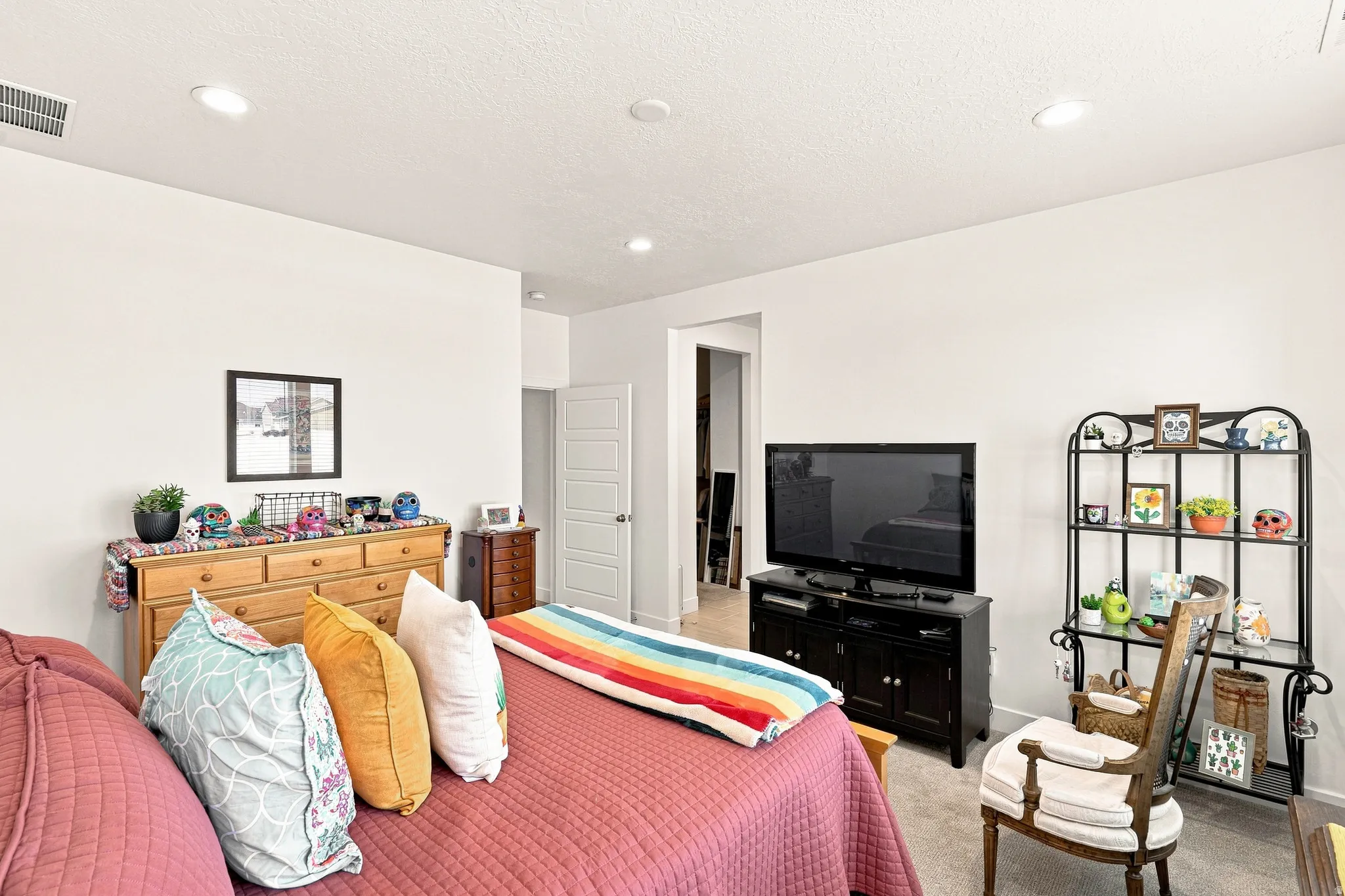 Spacious secondary bedroom featuring recessed ceiling lighting, wood-finish dresser with decorative hardware, wall-mounted flat-screen television, metal shelving unit with open storage, and carpet flooring throughout