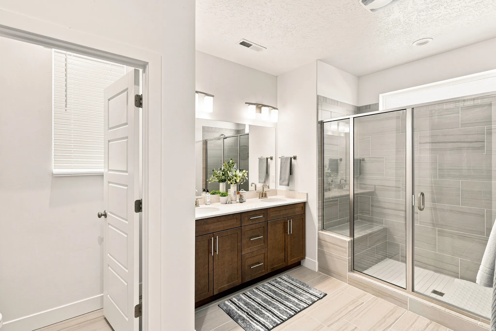 Spa-inspired primary bathroom featuring a wood-finish vanity with dual sinks, quartz countertop, and polished chrome hardware