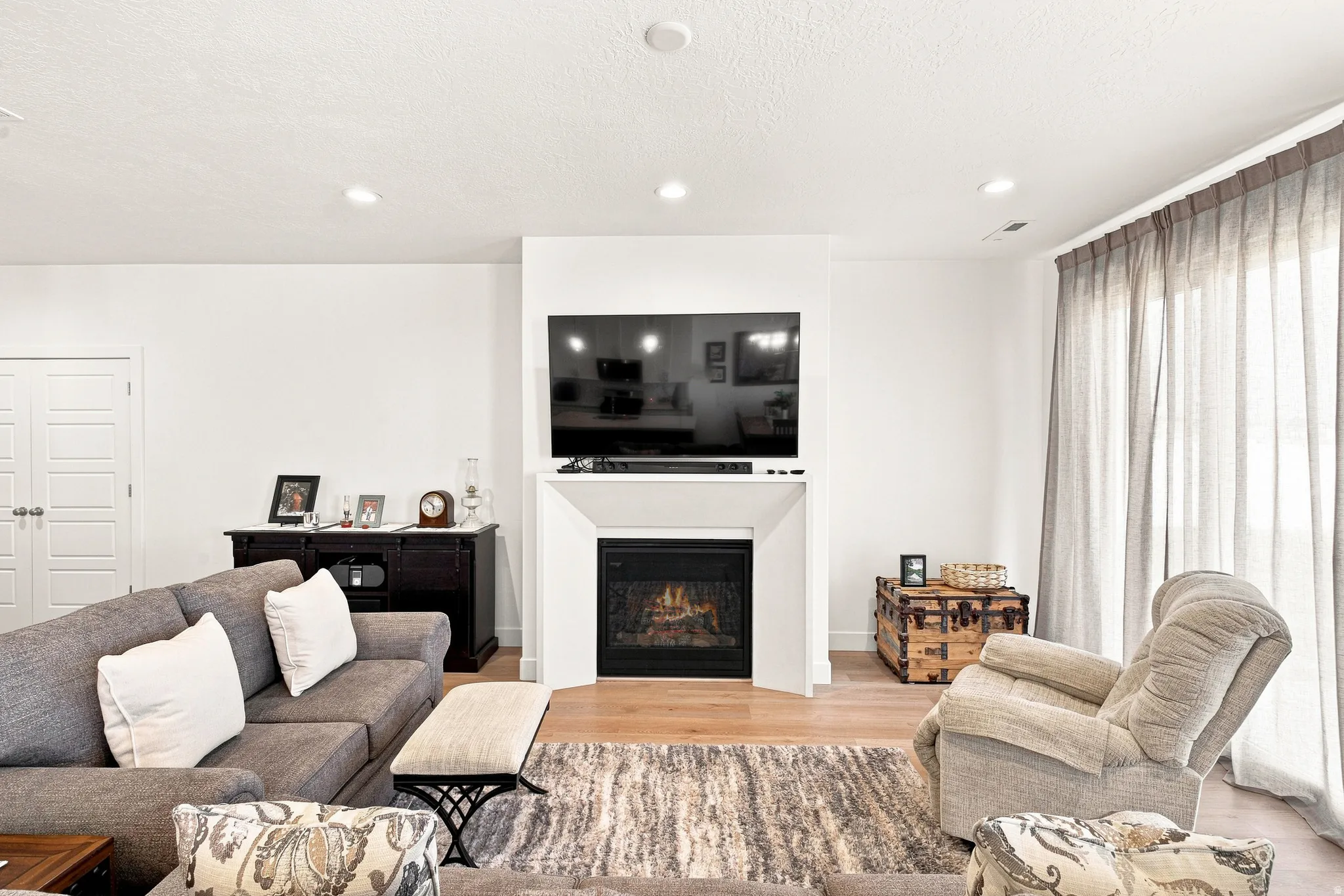 Bright, open living room featuring a gas fireplace with built-in cabinetry, wall-mounted flat-screen television, recessed ceiling lighting, and expansive windows with floor-to-ceiling drapery